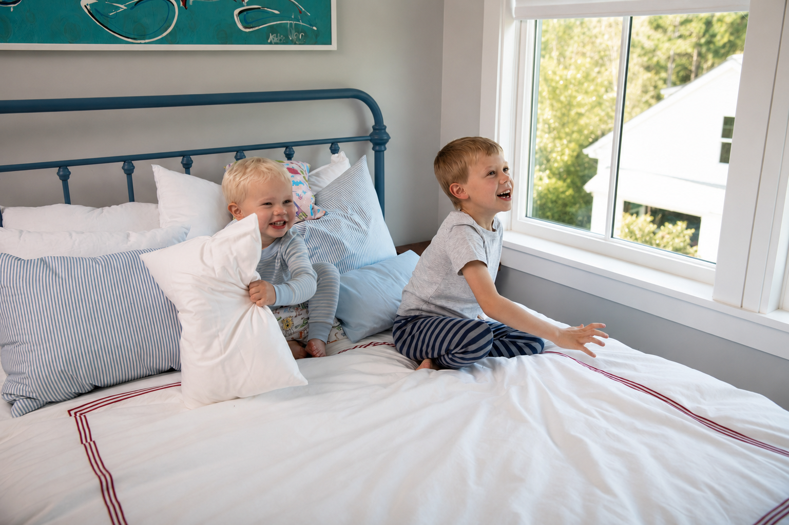 Two children playing on a white bed with striped pillows and a blue metal headboard in a bright bedroom, with a boy looking out a window.